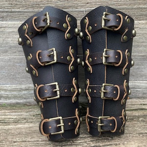 May include: A pair of black leather bracers featuring four horizontal straps with antique brass buckles. Each strap is secured to the main leather body with brass rivets. The leather has a visible grain texture and the edges are finished in a natural tan.