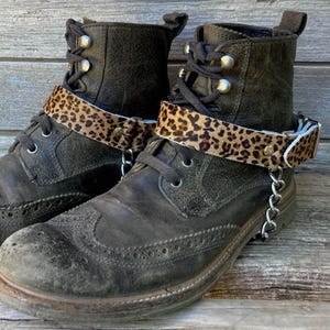 May include: A pair of worn, dark brown leather boots with decorative stitching and a leopard print ankle cuff. The cuffs are secured with small metal studs and a silver chain. The boots have laces and metal eyelets.