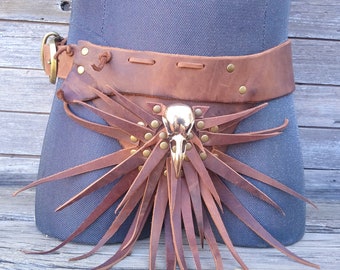 Op de afbeelding: Een bruine leren riem met een schedelvormige gesp en lange leren franjes. De riem is versierd met messing studs en klinknagels.