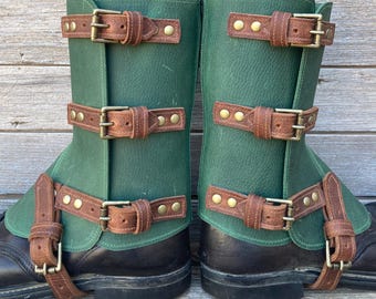 May include: A pair of green leather boot covers with brown leather straps and metal buckles. The boot covers are designed to fit over boots and have three adjustable straps. The boots are black.