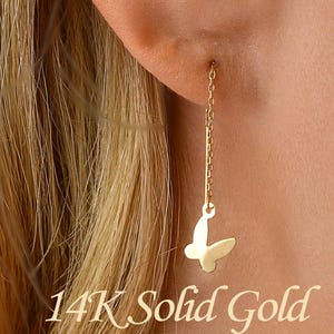 Op de afbeelding: Een gouden vlinder oorbellen. De oorbel hangt aan een fijne ketting. De tekst "14K Solid Gold" staat onderaan de afbeelding. De persoon heeft blond haar.