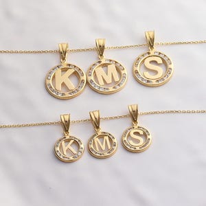 May include: Gold-toned initial pendant necklaces. Each pendant features a letter (K, M, or S) in a circular frame, embellished with small, clear stones. The pendants hang from delicate gold chains, creating a personalized jewelry set.