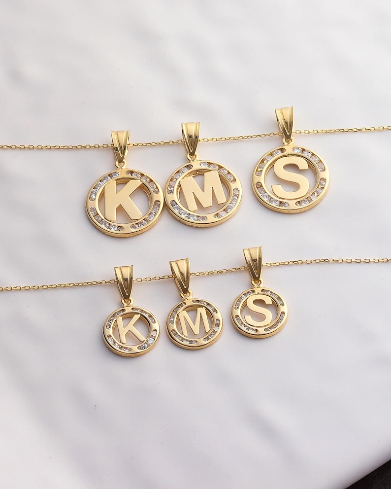 May include: Gold-toned initial pendant necklaces. Each pendant features a letter (K, M, or S) in a circular frame, embellished with small, clear stones. The pendants hang from delicate gold chains, creating a personalized jewelry set.