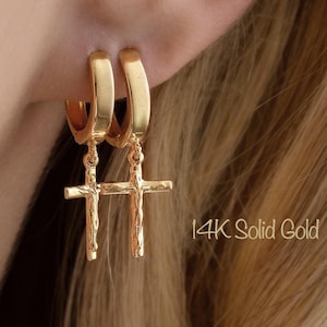 May include: Gold hoop earrings with cross pendants. The earrings are made of 14K solid gold. The text "14K Solid Gold" is visible. The earrings are shown on an ear.