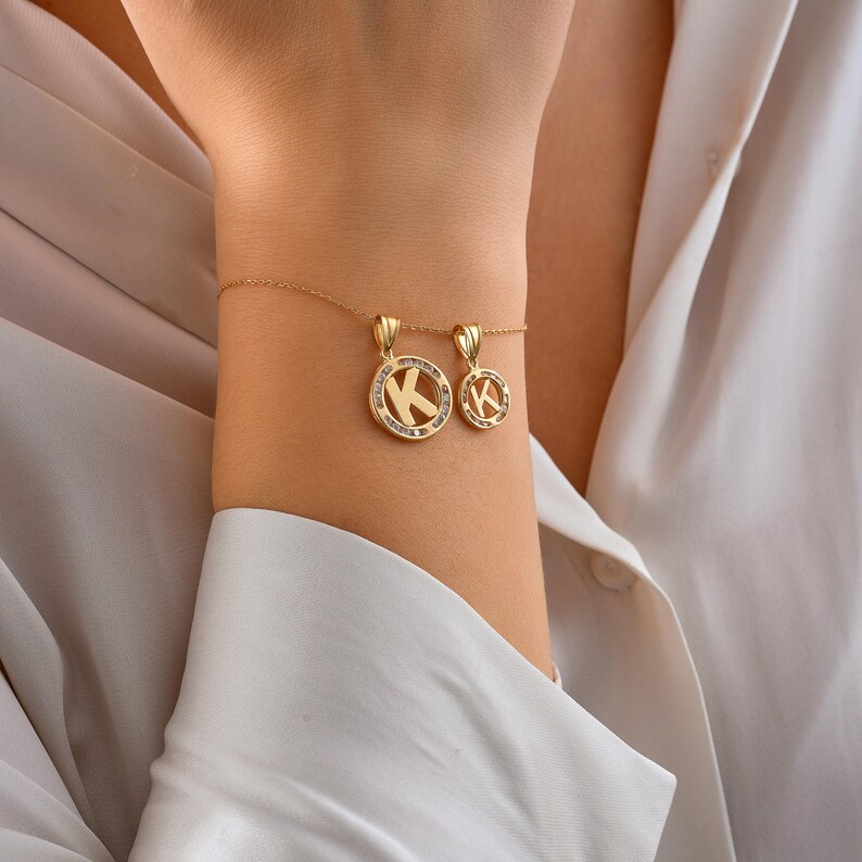 May include: A gold necklace and bracelet set featuring pendants with the letter "K". The pendants are circular, with the letter in the center and surrounded by small, clear stones. The jewelry is displayed on a person's wrist.