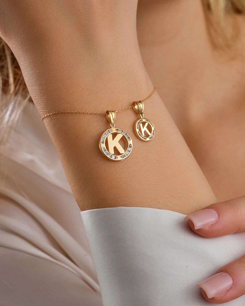 May include: A gold-colored bracelet with two pendants. Each pendant features the letter "K" in a circular design. The larger pendant is surrounded by small, clear stones. The bracelet is on a wrist.