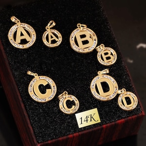 May include: Gold-colored initial pendants, featuring the letters A, B, C, and D, are displayed in a presentation box. Each pendant is encircled with small, clear gemstones. A small tag reads "14K".