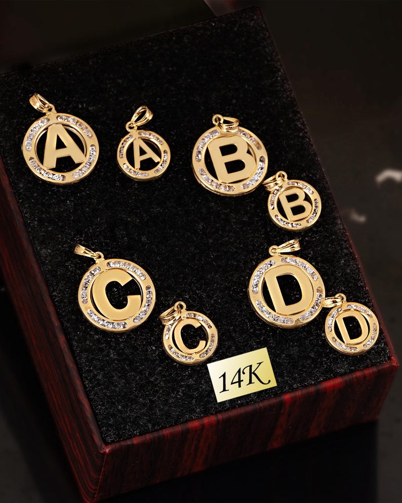 May include: Gold-colored initial pendants, featuring the letters A, B, C, and D, are displayed in a presentation box. Each pendant is encircled with small, clear gemstones. A small tag reads "14K".