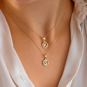 May include: Two gold necklaces with pendant charms. The top necklace features a pendant with the letter "K" in a circular design, and the bottom necklace has a pendant with the letter "S". Both pendants are surrounded by small, sparkling accents.