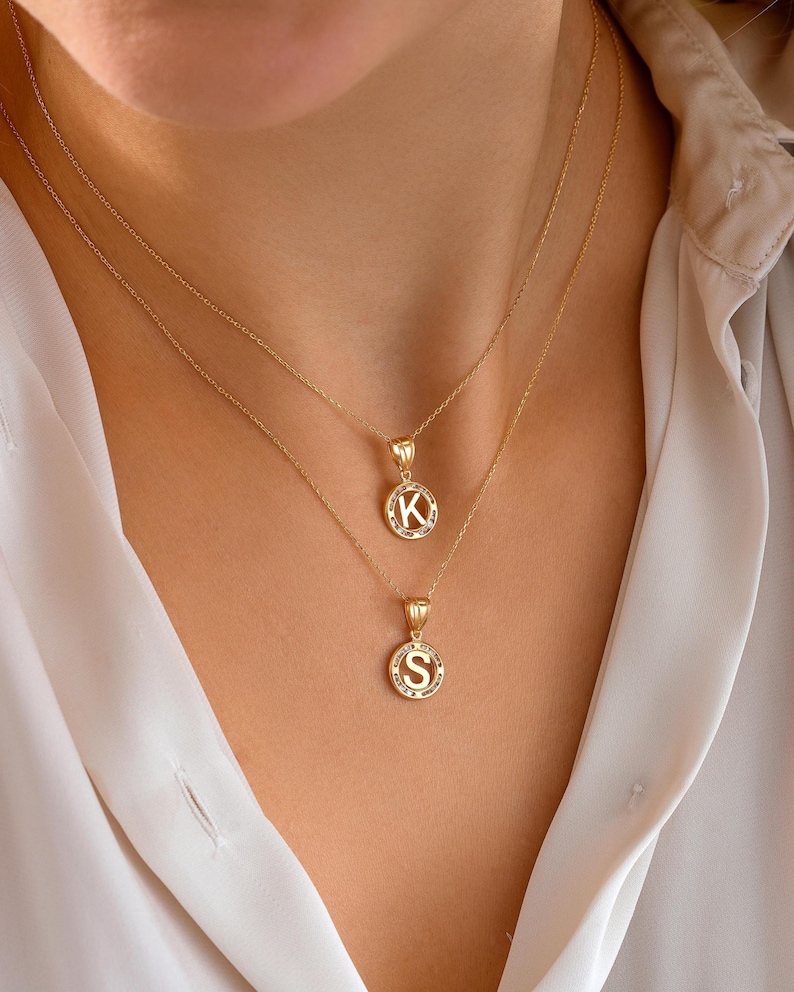 May include: Two gold necklaces with pendant charms. The top necklace features a pendant with the letter "K" in a circular design, and the bottom necklace has a pendant with the letter "S". Both pendants are surrounded by small, sparkling accents.