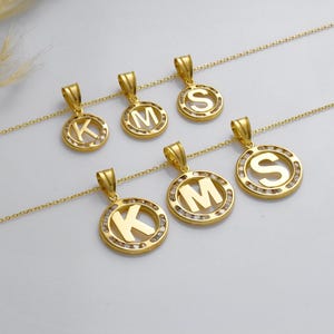 May include: Gold-colored initial pendant necklaces. Each necklace features a circular pendant with a letter (K, M, or S) in the center, surrounded by small, clear stones. The pendants hang from delicate gold chains.