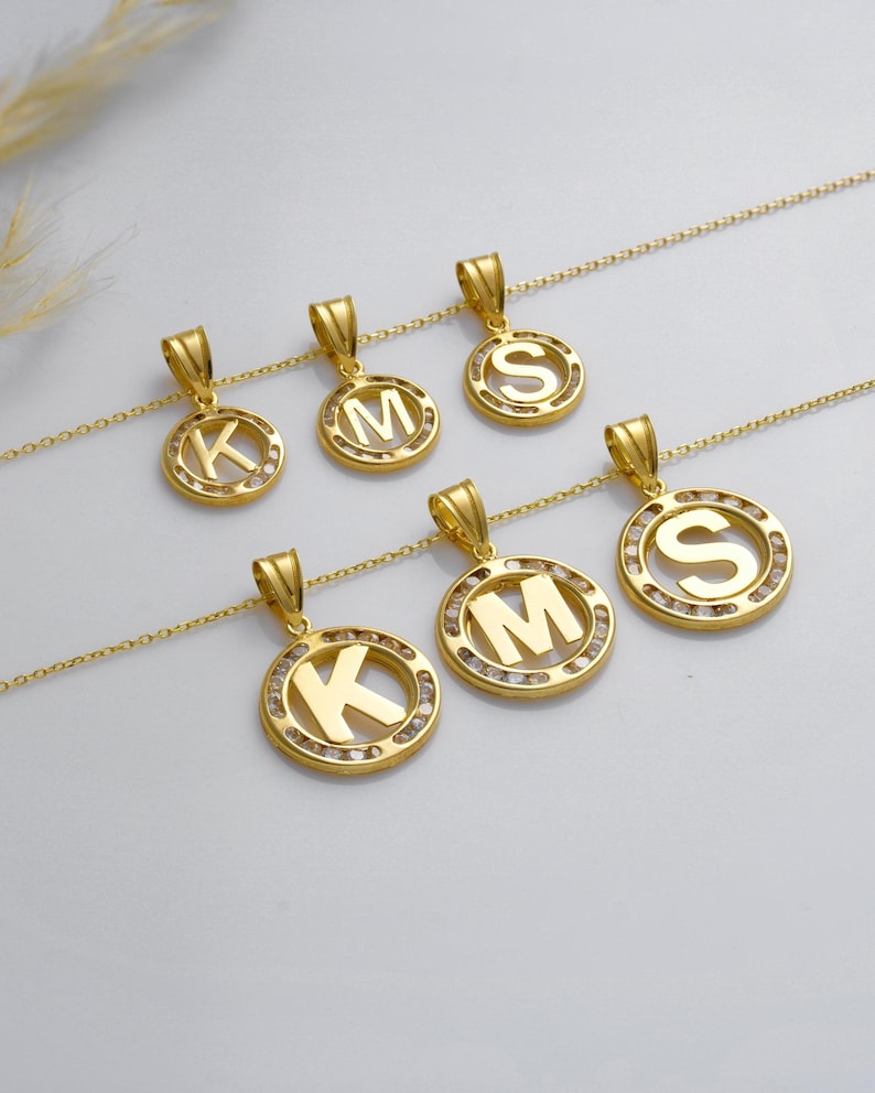 May include: Gold-colored initial pendant necklaces. Each necklace features a circular pendant with a letter (K, M, or S) in the center, surrounded by small, clear stones. The pendants hang from delicate gold chains.