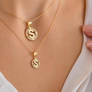 May include: Two gold necklaces with initial pendants. The pendants are circular, with the letter "S" in the center, surrounded by small, clear stones. The necklaces are displayed on a person wearing a white shirt.