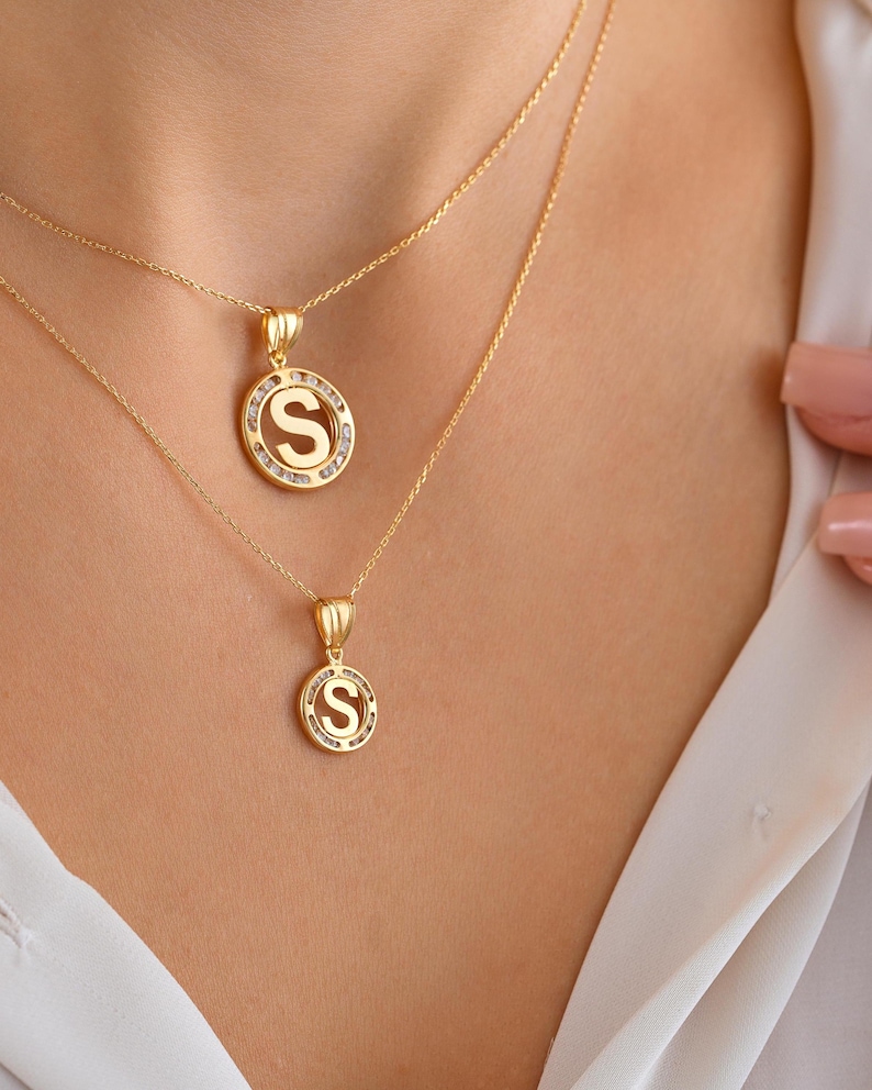 May include: Two gold necklaces with initial pendants. The pendants are circular, with the letter "S" in the center, surrounded by small, clear stones. The necklaces are displayed on a person wearing a white shirt.