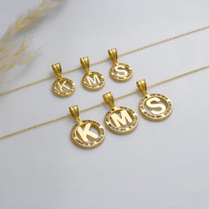 May include: Gold-colored initial pendant necklaces. Each necklace features a circular pendant with a letter in the center, surrounded by small, clear stones. The letters visible are K, M, and S. The pendants hang from delicate gold chains.