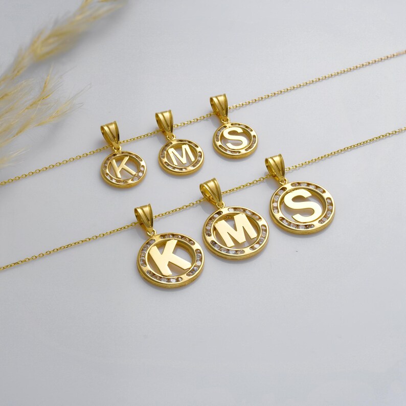 May include: Gold-colored initial pendant necklaces. Each necklace features a circular pendant with a letter in the center, surrounded by small, clear stones. The letters visible are K, M, and S. The pendants hang from delicate gold chains.