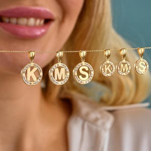 May include: Gold-colored initial pendants with the letters K, M, and S. Each pendant is circular, with a letter in the center and surrounded by small, clear stones. The pendants hang from delicate gold chains.