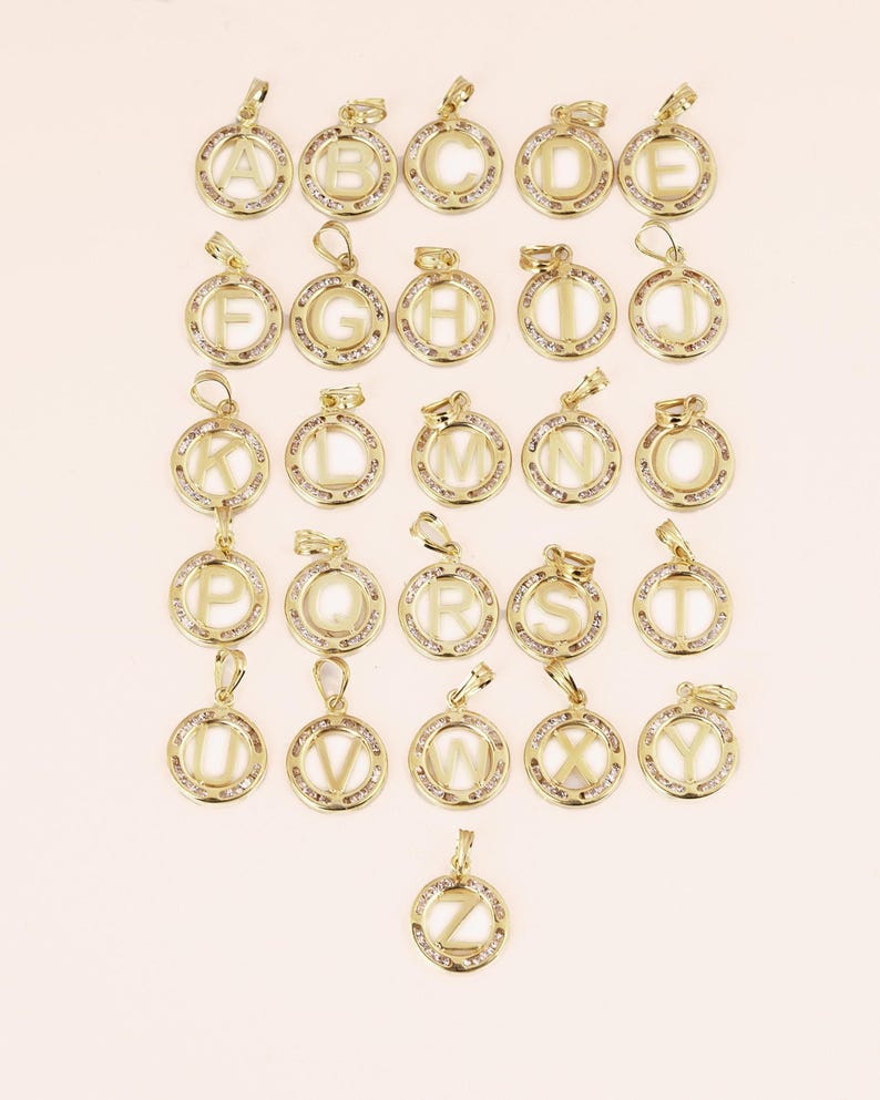 May include: A collection of gold-colored alphabet pendants, each featuring a letter from A to Z. The pendants are round, with a clear center and a border of small, sparkling stones. Each pendant has a loop for a chain.