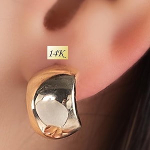 Puede incluir: Primer plano de un pendiente dorado con un diseño curvo y pulido. El pendiente está marcado con "14K". El pendiente es de estilo media argolla, reflejando la luz y añadiendo un toque de elegancia. El pendiente se lleva en una oreja.