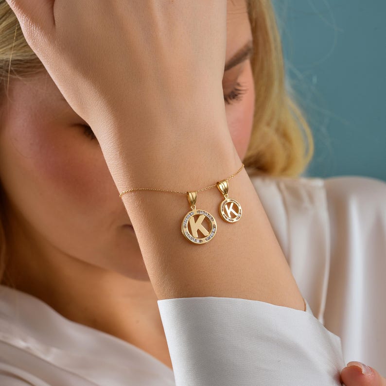 May include: A gold-colored necklace and bracelet set featuring the letter "K" in a pendant design. The larger pendant is encircled with small, clear gemstones. The jewelry is displayed on a person's arm, showcasing the delicate chain and pendant details.
