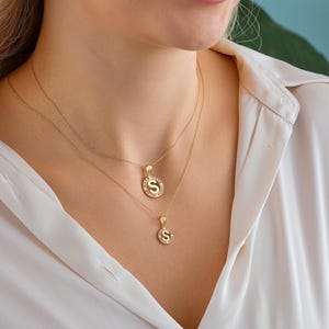 May include: Two gold necklaces with pendant charms. The top necklace has a pendant with the letter "S" surrounded by small clear stones. The second necklace has a smaller pendant with the letter "S". The necklaces are worn over a white blouse.