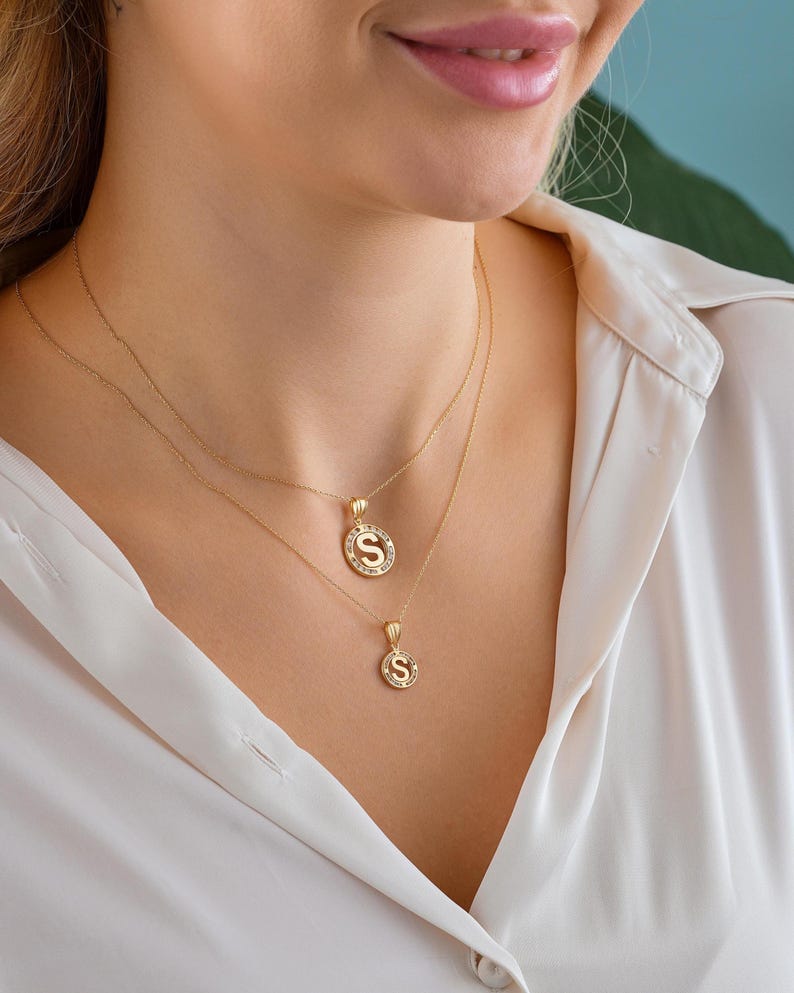 May include: Two gold necklaces with pendant charms. The top necklace has a pendant with the letter "S" surrounded by small clear stones. The second necklace has a smaller pendant with the letter "S". The necklaces are worn over a white blouse.