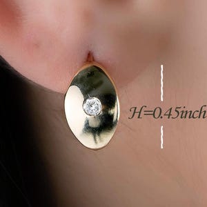 May include: A close-up of a gold earring with a small, round, clear gemstone in the center. The oval-shaped earring is 1.14 cm in height. The earring is set against a blurred background.