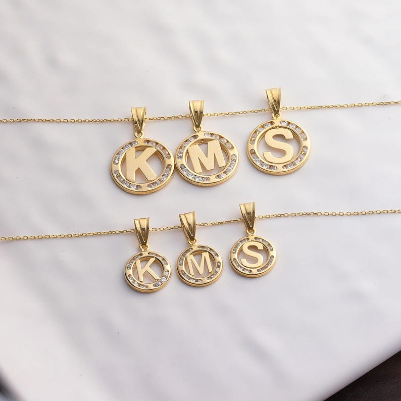 May include: Gold-colored initial pendant necklaces. Each pendant features a letter (K, M, or S) within a circular frame, embellished with small, clear stones. The pendants hang from delicate gold chains, creating a personalized jewelry set.