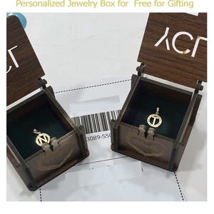 May include: Two wooden jewelry boxes with open lids, each containing a gold pendant with a letter. The boxes are brown with dark green velvet lining. The text "Personalized Jewelry Box for Free for Gifting" is at the top.
