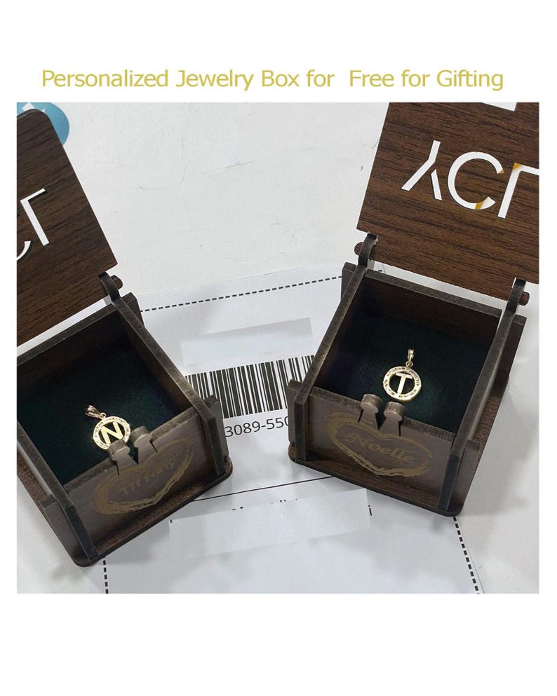 May include: Two wooden jewelry boxes with open lids, each containing a gold pendant with a letter. The boxes are brown with dark green velvet lining. The text "Personalized Jewelry Box for Free for Gifting" is at the top.