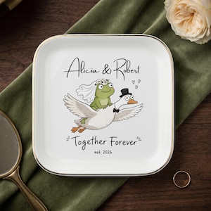 May include: A square white ceramic trinket dish with a gold-toned rim featuring a cartoon illustration of a frog in a wedding veil riding a goose wearing a top hat and bowtie. The text reads Alicia & Robert, Together Forever, and est. 2026.