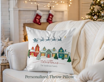 May include: White throw pillow with a Christmas village scene. The design features a sleigh pulled by reindeer above colorful houses and the text "Merry Christmas" and "The Sanderson Family". The pillow is on a white couch.