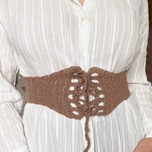 May include: A brown crocheted corset belt with a lace-up front. The belt is tied in a bow at the front.