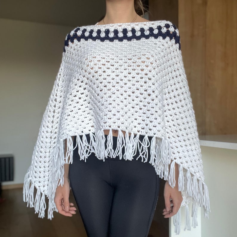 Puede incluir: Un poncho blanco de ganchillo con un ribete azul marino en el escote. El poncho presenta flecos en el borde inferior. El dise&ntilde;o de tejido abierto es visible en toda la prenda, creando un aspecto ligero y aireado. Este accesorio hecho a mano.