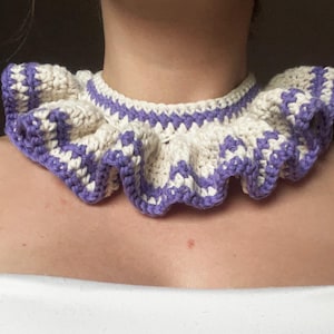 Può includere: Un colletto all'uncinetto color crema e lavanda. Il colletto presenta un bordo a balze e un motivo a zig-zag in lavanda. Il colletto è indossato attorno al collo ed è realizzato in filato morbido.