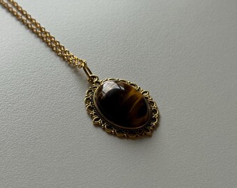 May include: A gold-colored necklace featuring an oval pendant with a tiger's eye stone. The pendant is set in a decorative, filigree-style frame. The chain is delicate and also gold-colored, creating a vintage aesthetic.