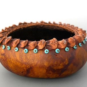 May include: A round, brown gourd bowl featuring a textured, dark brown bark-like rim. The exterior is decorated with a single row of small, round, light blue beads attached with thin brown cord, encircling the upper portion of the bowl.