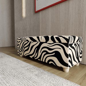 Könnte beinhalten: Eine rechteckige Aufbewahrungs-Ottomane mit einem auffälligen, schwarz-cremefarbenen, wellenförmigen Muster im Zebra-Stil. Das Möbelstück ist mit Stoff bezogen und steht auf vier kleinen, hellen Holzfüßen, was eine niedrige Sitz- oder Stauraumlösung bietet.