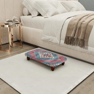 Puede incluir: Un pequeño reposapiés decorativo con una parte superior rectangular y cuatro patas cortas de madera. La parte superior presenta un diseño estampado colorido con tonos de rosa, turquesa y blanco. El reposapiés está colocado sobre una alfombra blanca cerca de una cama.