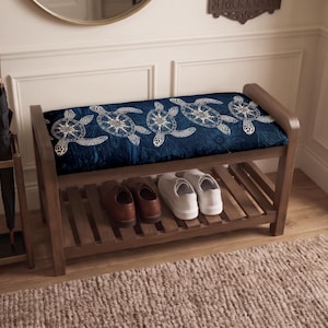 May include: A wooden entryway bench featuring a cushioned seat upholstered in a dark blue fabric with a repeating white sea turtle pattern. The bench includes a lower slatted shelf for storage, currently holding a pair of brown shoes and white sneakers.