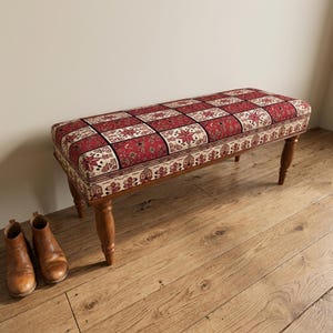 Peut inclure: Un banc rectangulaire rembourré avec un motif patchwork rouge et crème orné de détails floraux et géométriques. Le banc repose sur quatre pieds en bois tourné de couleur brun moyen. Deux bottines en cuir marron sont posées à côté.