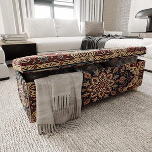 May include: A patterned storage ottoman with a hinged lid, in red, blue, and gold. A light grey blanket with fringe detail is draped over the edge. The ottoman is on a light-coloured rug. The ottoman is a stylish piece of furniture.