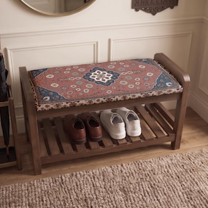 May include: A wooden shoe bench with a rectangular seat cushion featuring a red and blue intricate floral pattern. The bench has a lower slatted shelf holding a pair of brown leather shoes and a pair of white sneakers, positioned next to an umbrella stand.