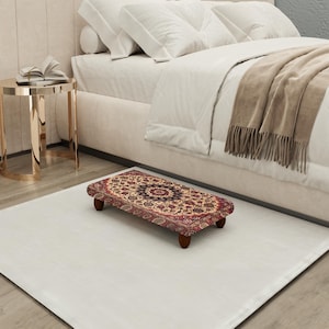 Puede incluir: Un pequeño reposapiés rectangular con una parte superior de alfombra estampada en tonos rojos, beige y marrón. El reposapiés tiene cuatro patas cortas de madera oscura y está colocado sobre una alfombra blanca cerca de una cama.