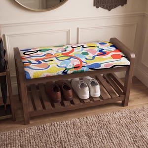 May include: A wooden shoe bench with a cushioned seat featuring a colorful abstract pattern of red, blue, yellow, and pink shapes on a white background. The bench has a slatted lower shelf holding a pair of brown leather shoes and white sneakers.