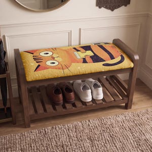 May include: A wooden shoe bench featuring a cushioned seat with a graphic illustration of an orange cat holding a cup. The bench has a slatted lower shelf holding a pair of brown leather shoes and a pair of white sneakers, with a wooden umbrella stand.