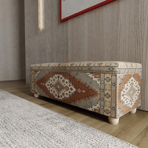 Puede incluir: Un otomán de almacenamiento rectangular con un diseño de tela estampada en tonos marrón, naranja y verde azulado. El otomán se asienta sobre cuatro patas cortas de madera clara. El diseño presenta formas geométricas y motivos tradicionales.