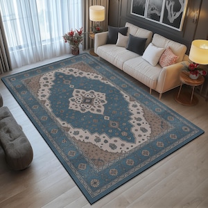 Könnte beinhalten: Ein rechteckiger Teppich mit einem aufwendigen, traditionellen Design. Der Teppich hat ein zentrales Medaillon in Blau-, Beige- und Brauntönen, umgeben von einem detaillierten Rand. Er liegt auf einem Holzboden, mit einem Sofa und einem Fenster im Hintergrund.