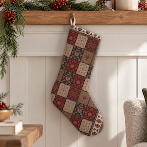 Puede incluir: Un calcetín navideño con un diseño de patchwork de patrones cuadrados alternos en tonos rojo intenso, verde oscuro y crema. La tela muestra intrincados motivos florales y geométricos, rematado con un borde decorativo en la parte superior.