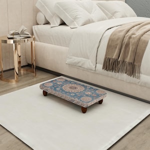 Puede incluir: Un pequeño reposapiés rectangular con una parte superior estampada en azul y beige y patas de madera oscura. El reposapiés está colocado sobre una alfombra blanca frente a una cama con ropa de cama blanca.